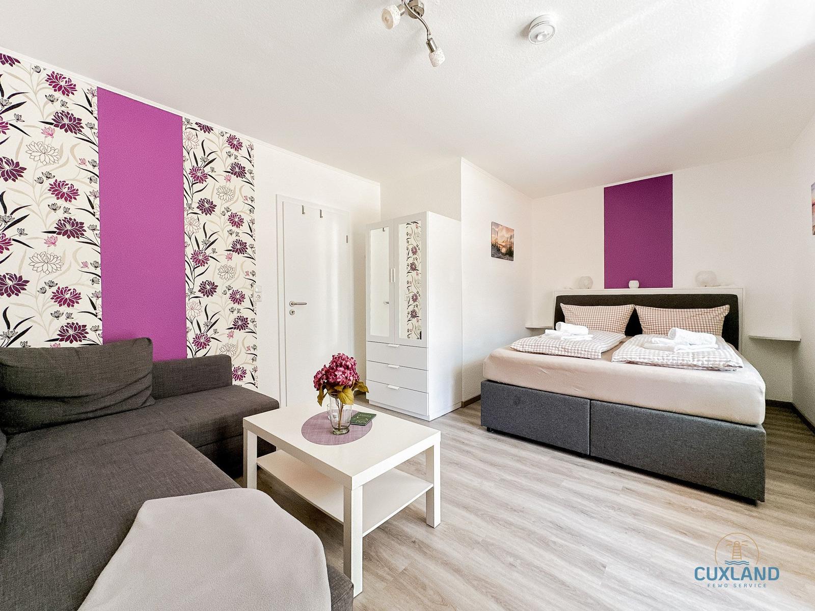 Schlafzimmer mit Bett, Schrank und Sofa. Wand mit floraler Tapete und lila Akzent.