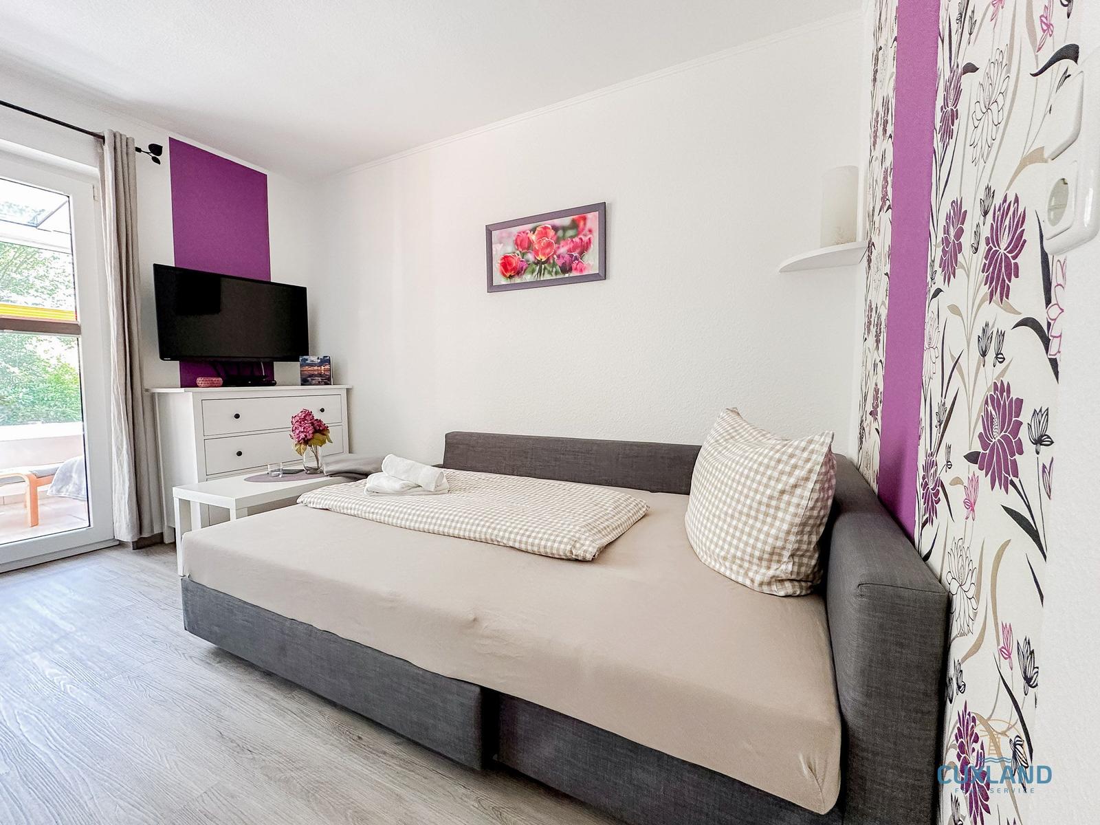 Schlafzimmer mit Bett, Fernseher und Balkontür.