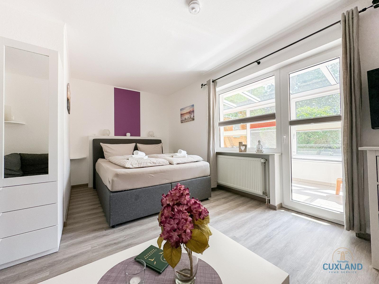 Schlafzimmer mit Doppelbett, Fenster und Balkonzugang. Möbel und Dekoration sind sichtbar.