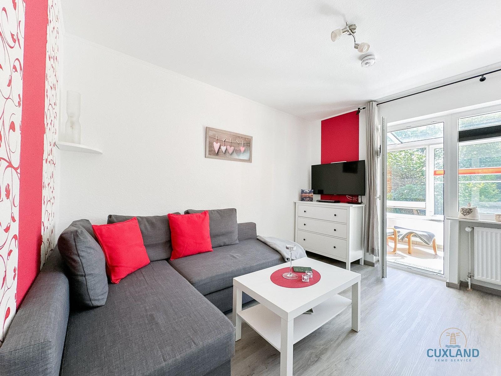 Wohnzimmer mit grauem Sofa, roten Kissen und weißem Couchtisch. Fernseher und Balkon mit Blick nach draußen.