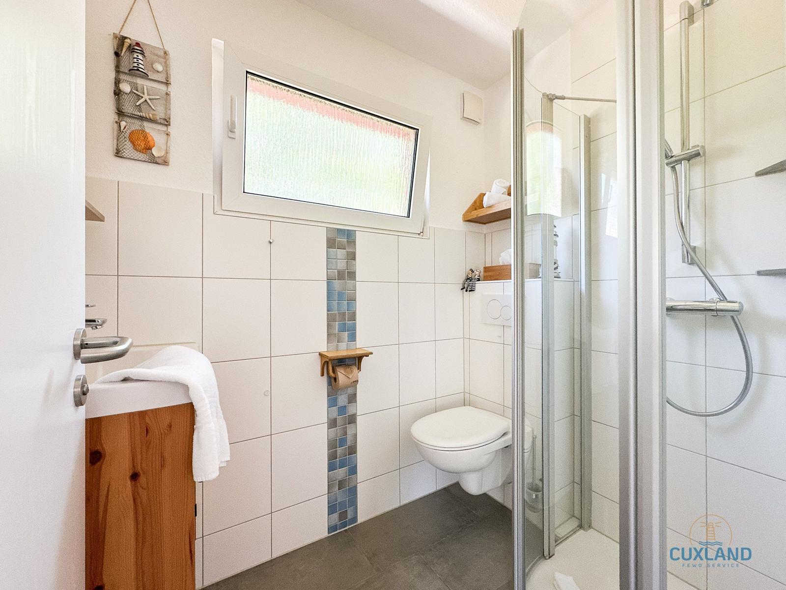 Badezimmer mit Dusche, WC und Waschbecken. Fenster mit Gitter und Holzregal.