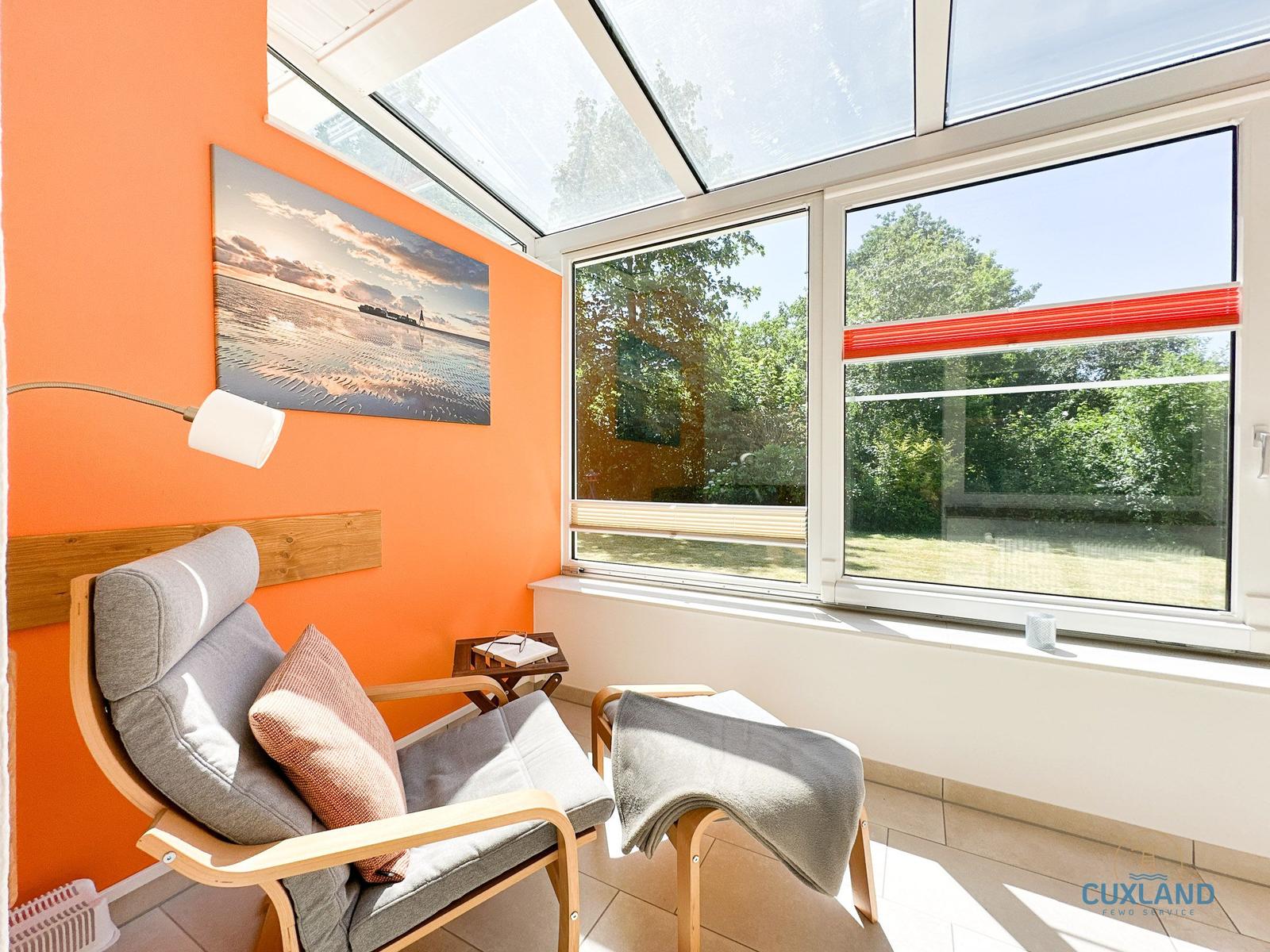 Raum mit orangefarbener Wand, Sitzmöbel und Fenster zur grünen Außenseite.