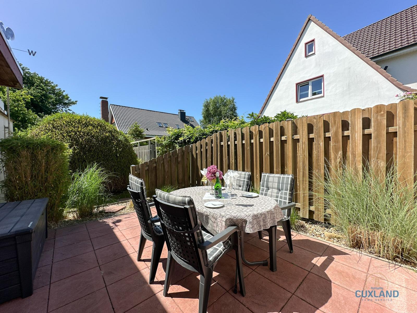 Terrasse mit Tisch und Stühlen, Holzzaun und Gartenpflanzen im Hintergrund.