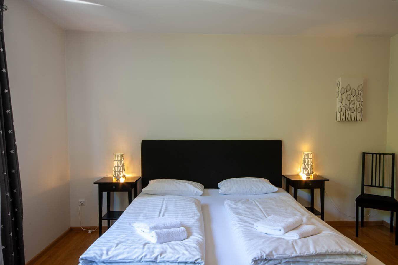 Schlafzimmer mit Doppelbett, zwei Nachttischen und Stuhl. Wandbild und Vorhänge sind sichtbar.