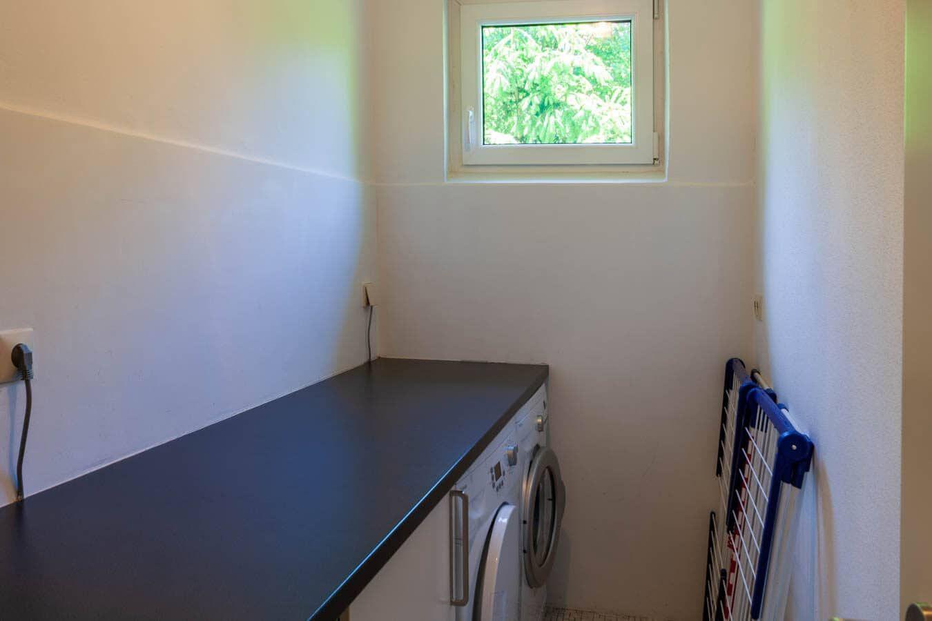 Wäscherei mit Waschmaschine, dunklem Arbeitsplatz und Fenster mit Blick auf Grün.