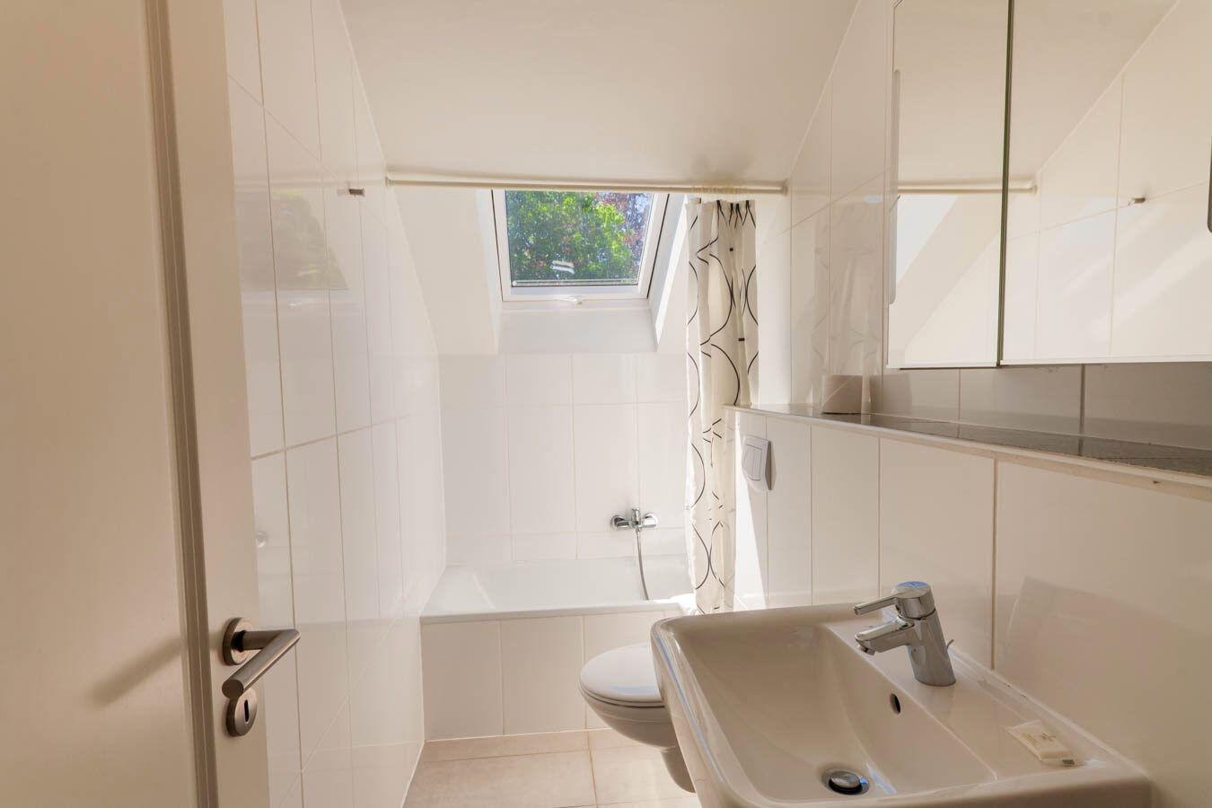 Badezimmer mit Dusche, Waschbecken und Fenster. Weiße Fliesen und Spiegel.
