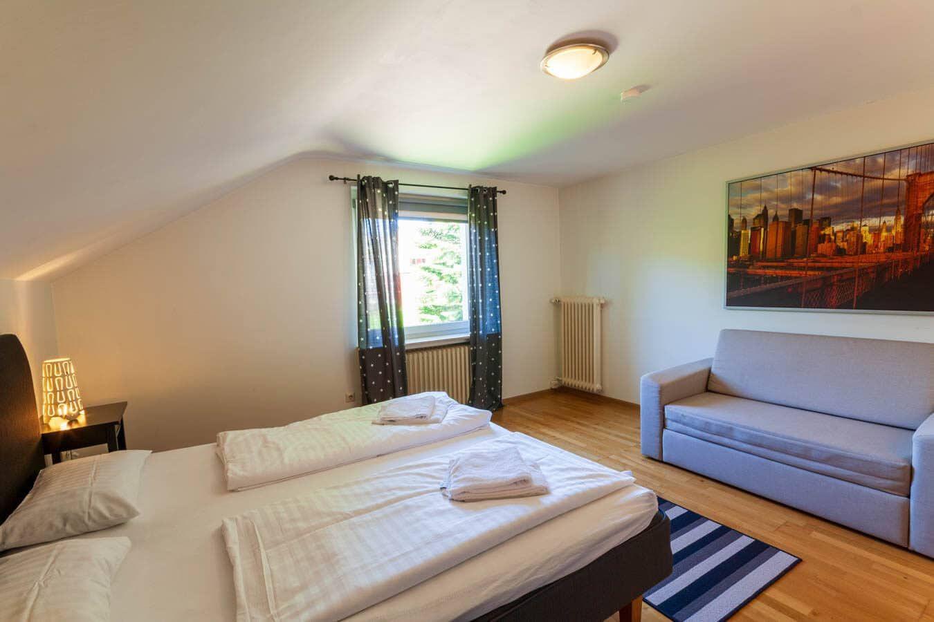 Zimmer mit Bett, Sofa und Fenster mit Blick. Wandbild zeigt Stadtansicht.