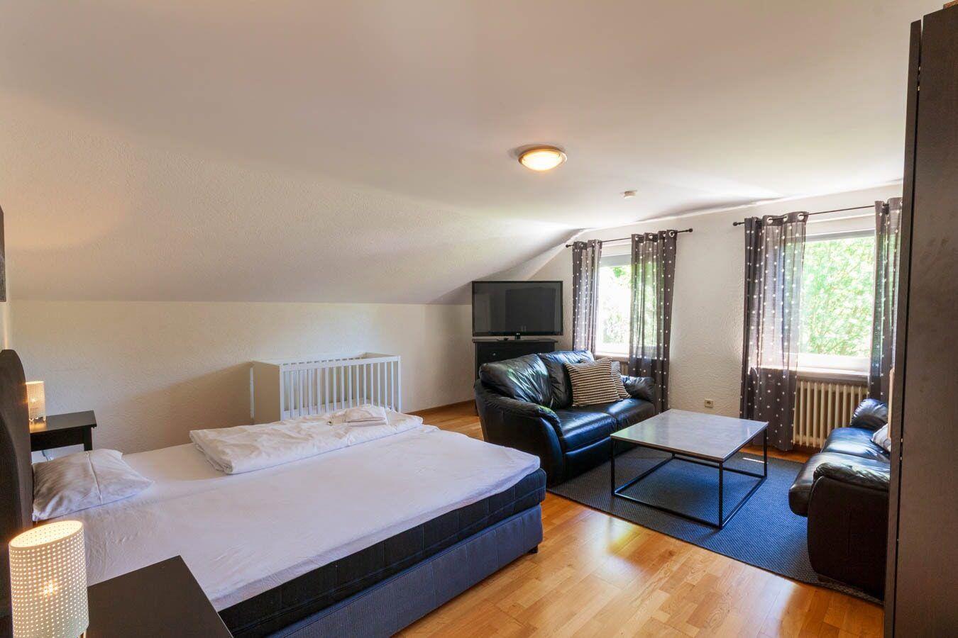 Schlafzimmer mit Bett, Wohnbereich mit Sofa und Fernseher, Holzboden und Fenster mit Vorhängen.