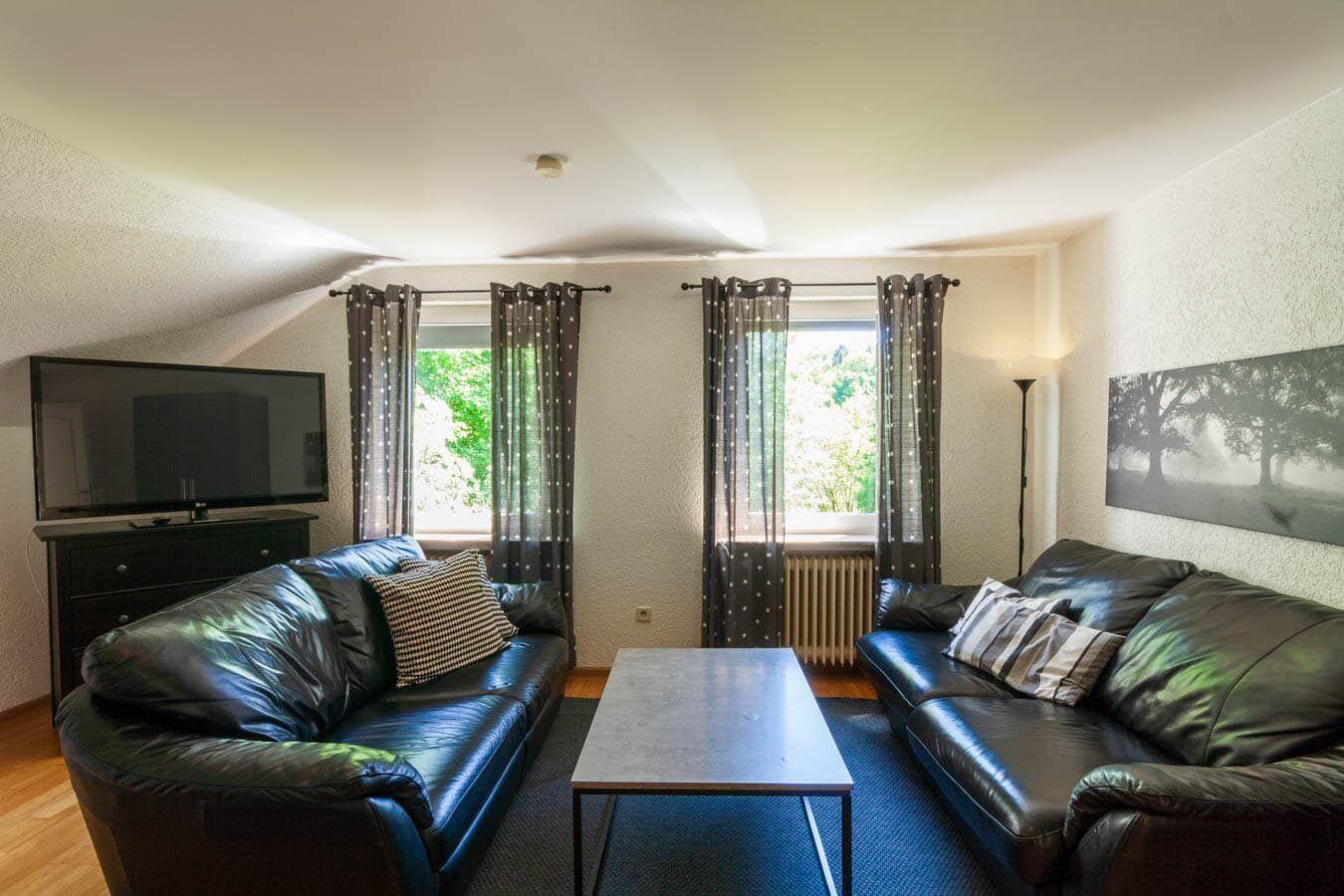 Wohnzimmer mit zwei Ledersofas, Fernseher und Fenstern mit Vorhängen.