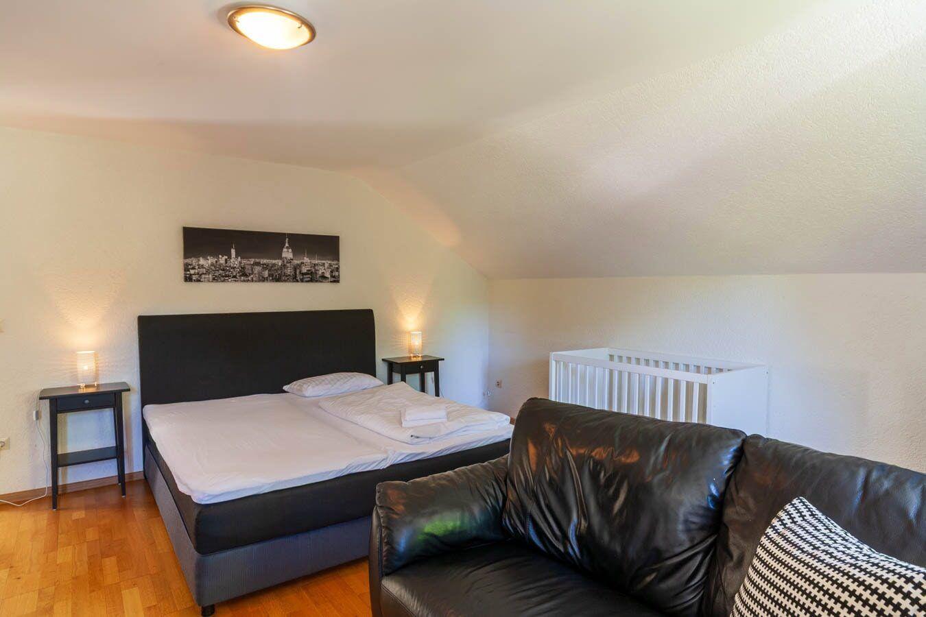 Schlafzimmer mit Doppelbett, Sofa und Wiege. Holzboden und schlichte Einrichtung.