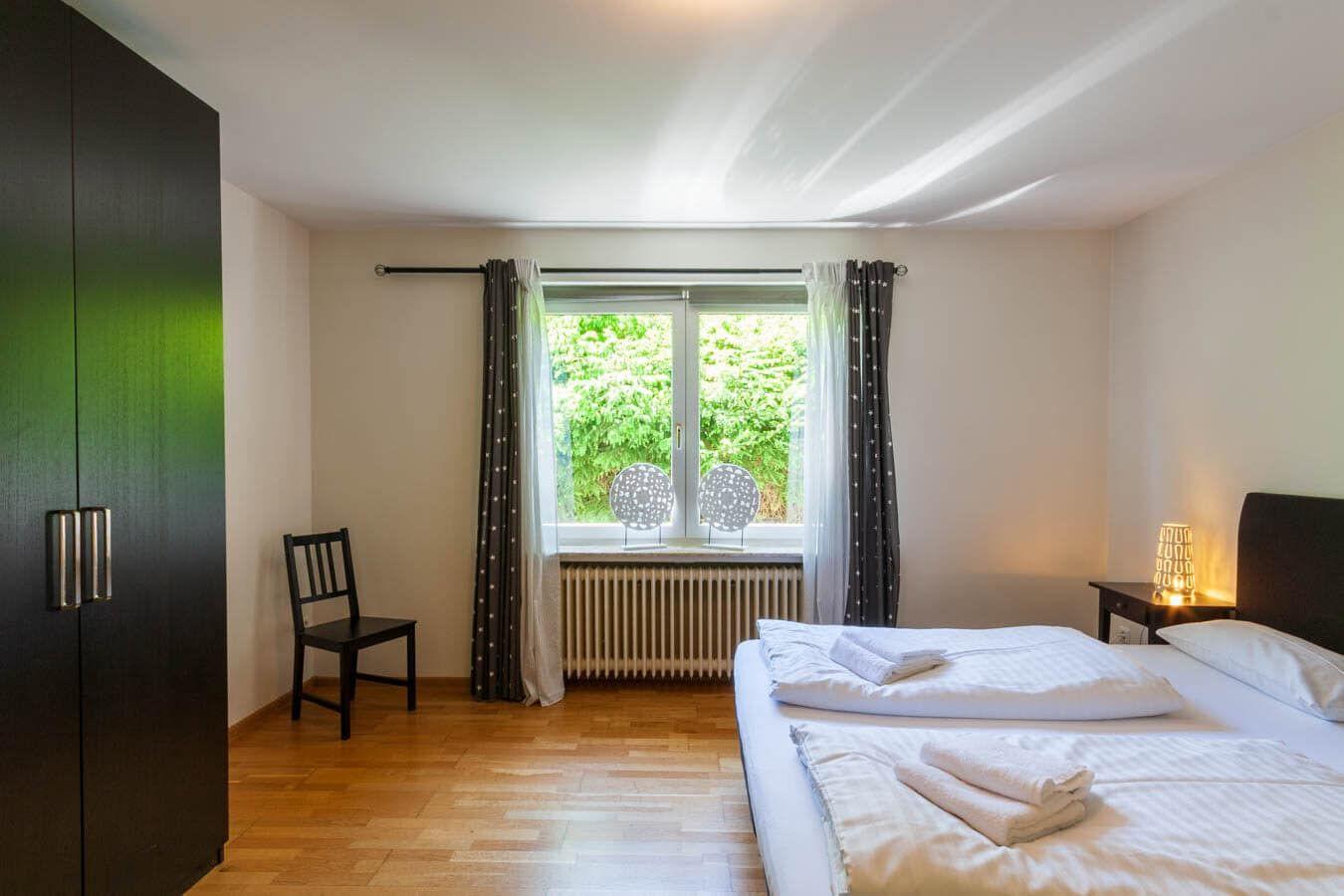 Schlafzimmer mit Bett, Kleiderschrank, Stuhl und Fenster mit Blick auf Grün.