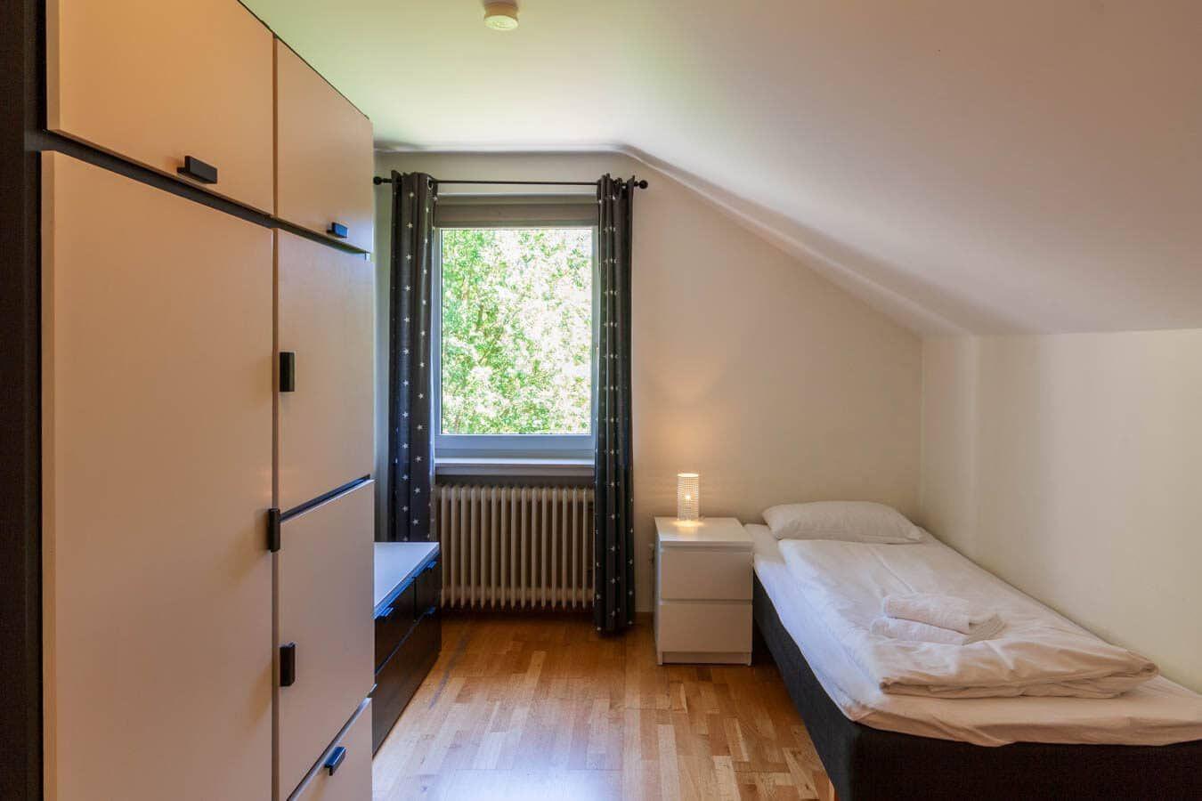 Schlafzimmer mit Einzelbett, Kleiderschrank und Fenster mit Blick auf Bäume.