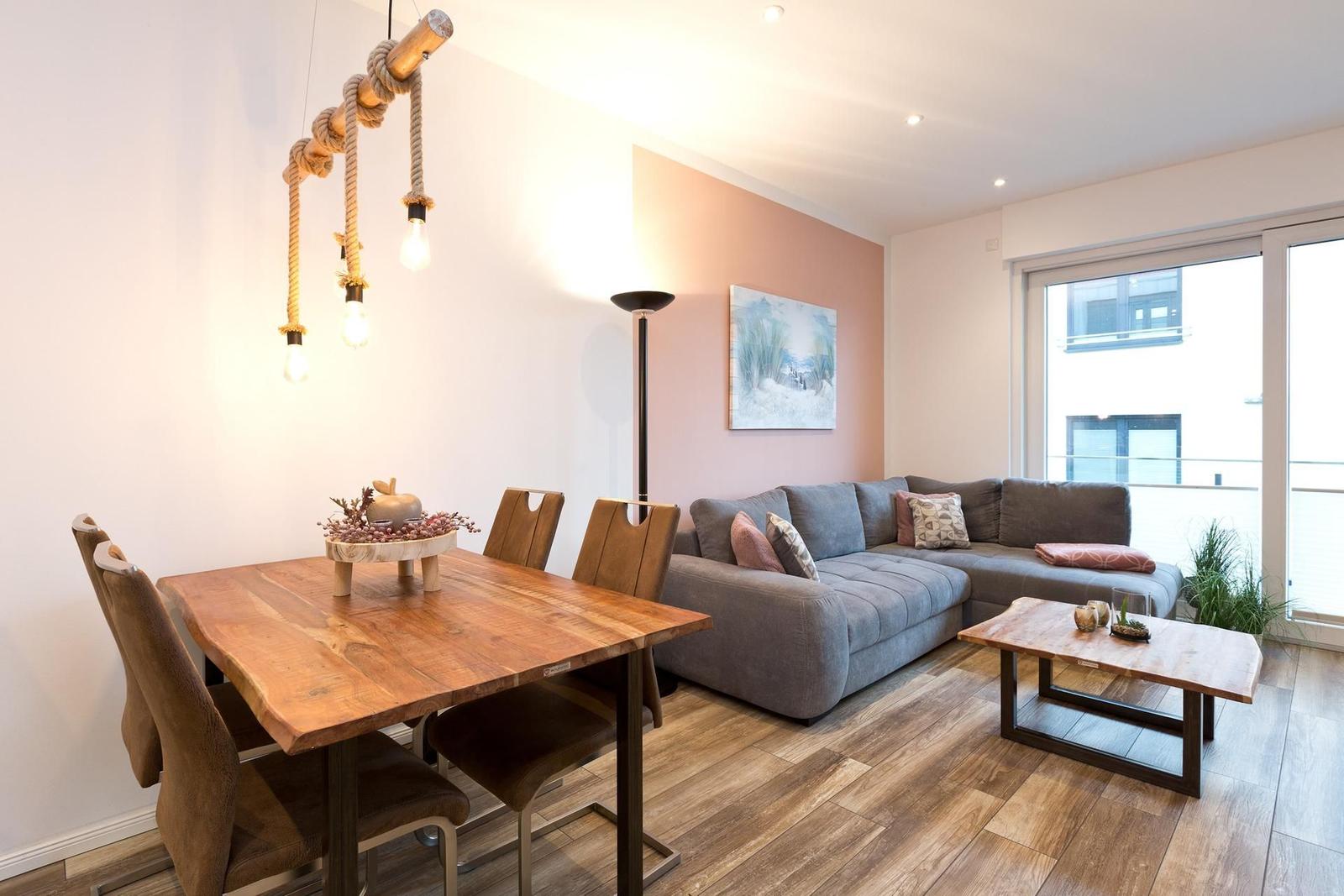 Essbereich mit Holztisch und Stühlen. Wohnbereich mit Sofa und Couchtisch. Holzdielenboden und große Fenster.