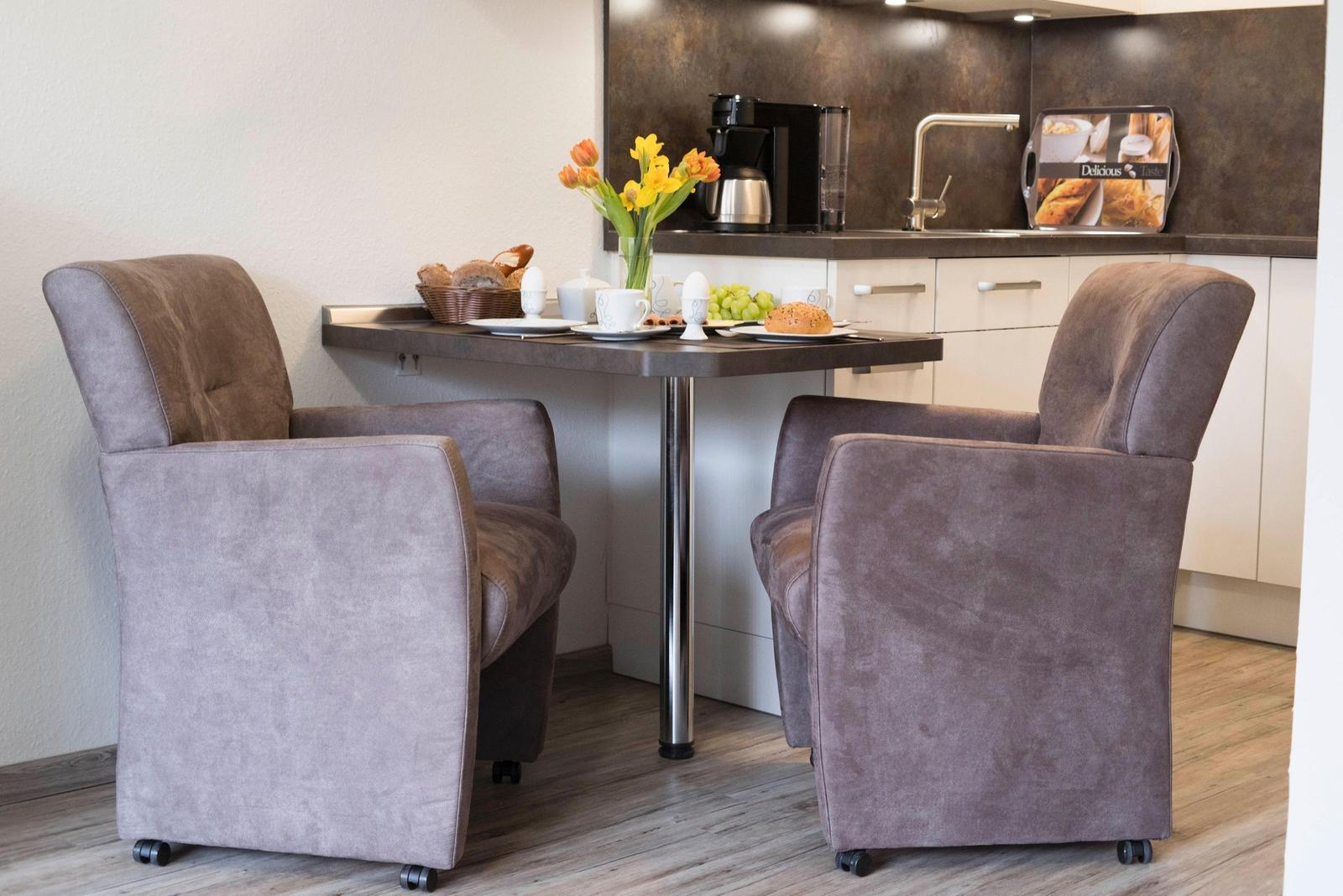 Zwei graue Sessel stehen an einem Tisch mit Frühstück und Blumen. Hintergrund ist Küche mit Arbeitsplatte.