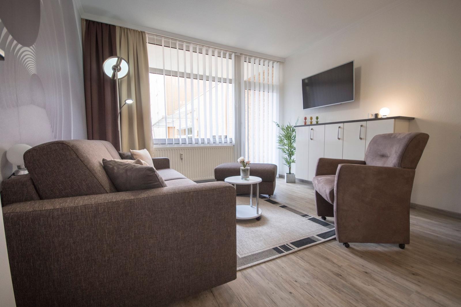 Wohnzimmer mit Sofa, Sessel, Fernseher und Fenster mit Jalousien.