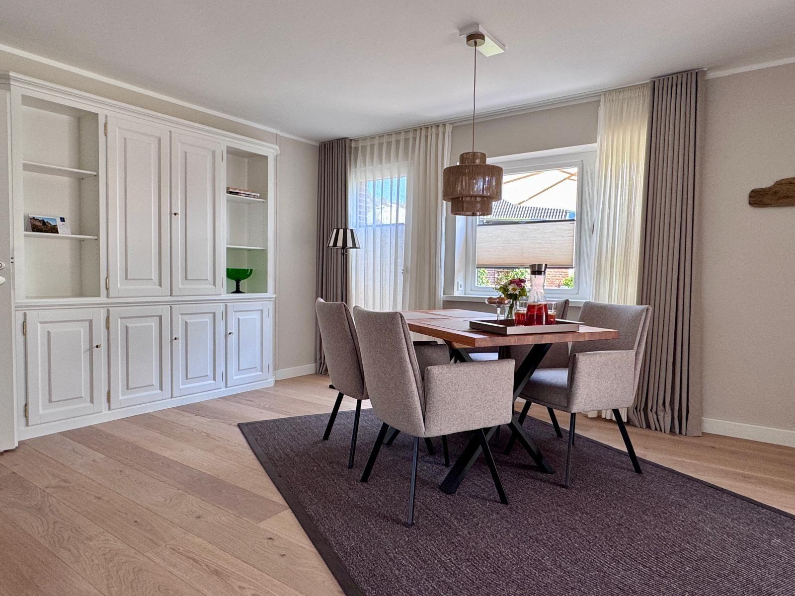 Esszimmer mit Holztisch, Stühlen und weißen Schränken. Fenster mit Vorhängen und Deckenleuchte.