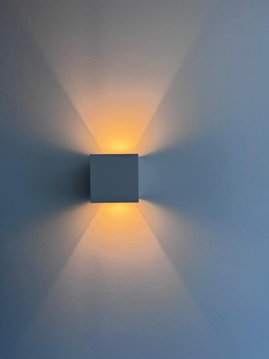 Eine quadratische Wandlampe mit warmweißem Licht an einer hellen Wand.
