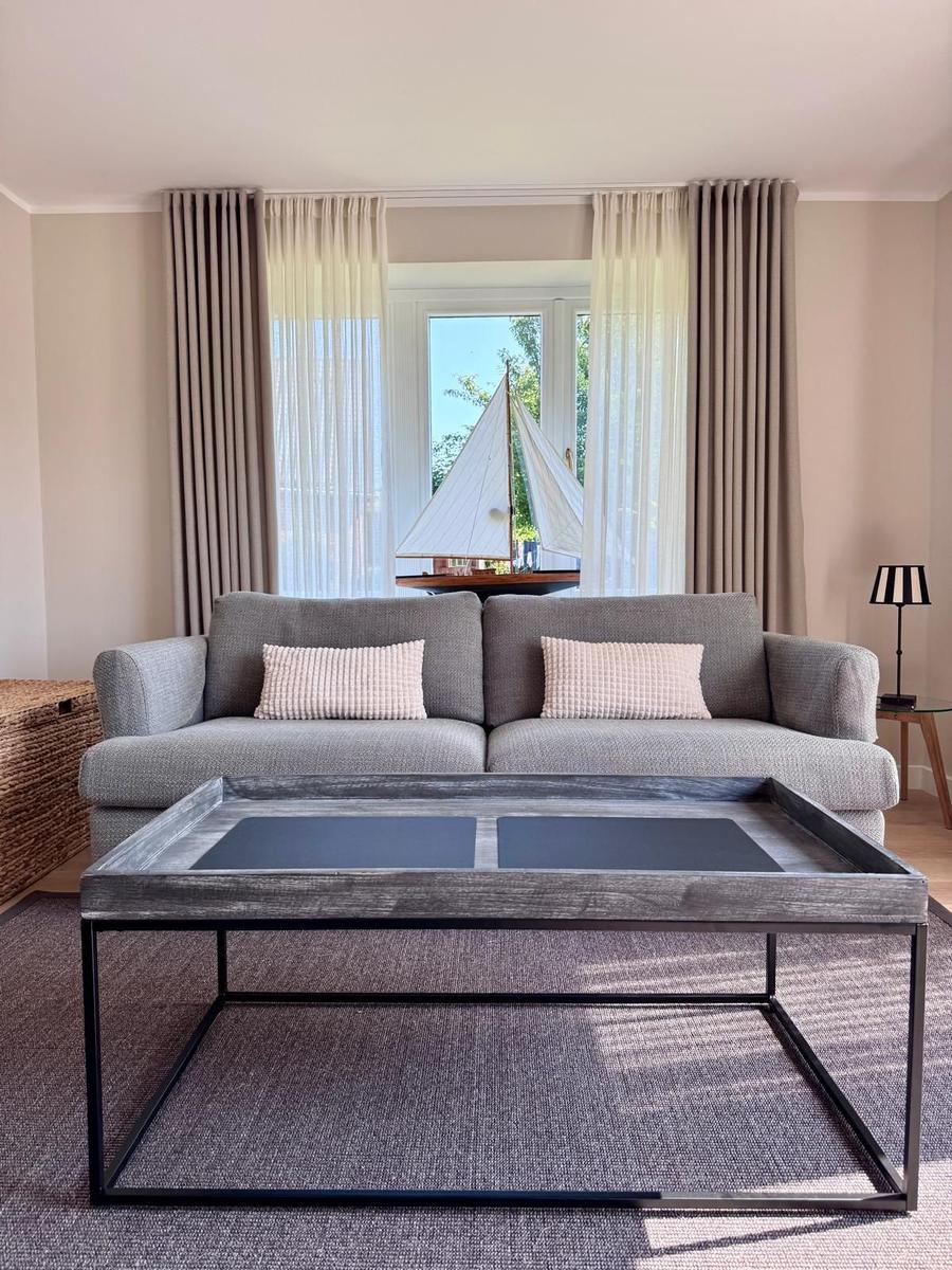 Ein Wohnzimmer mit grauem Sofa, Tisch und Fenster mit Segelbootmodell.