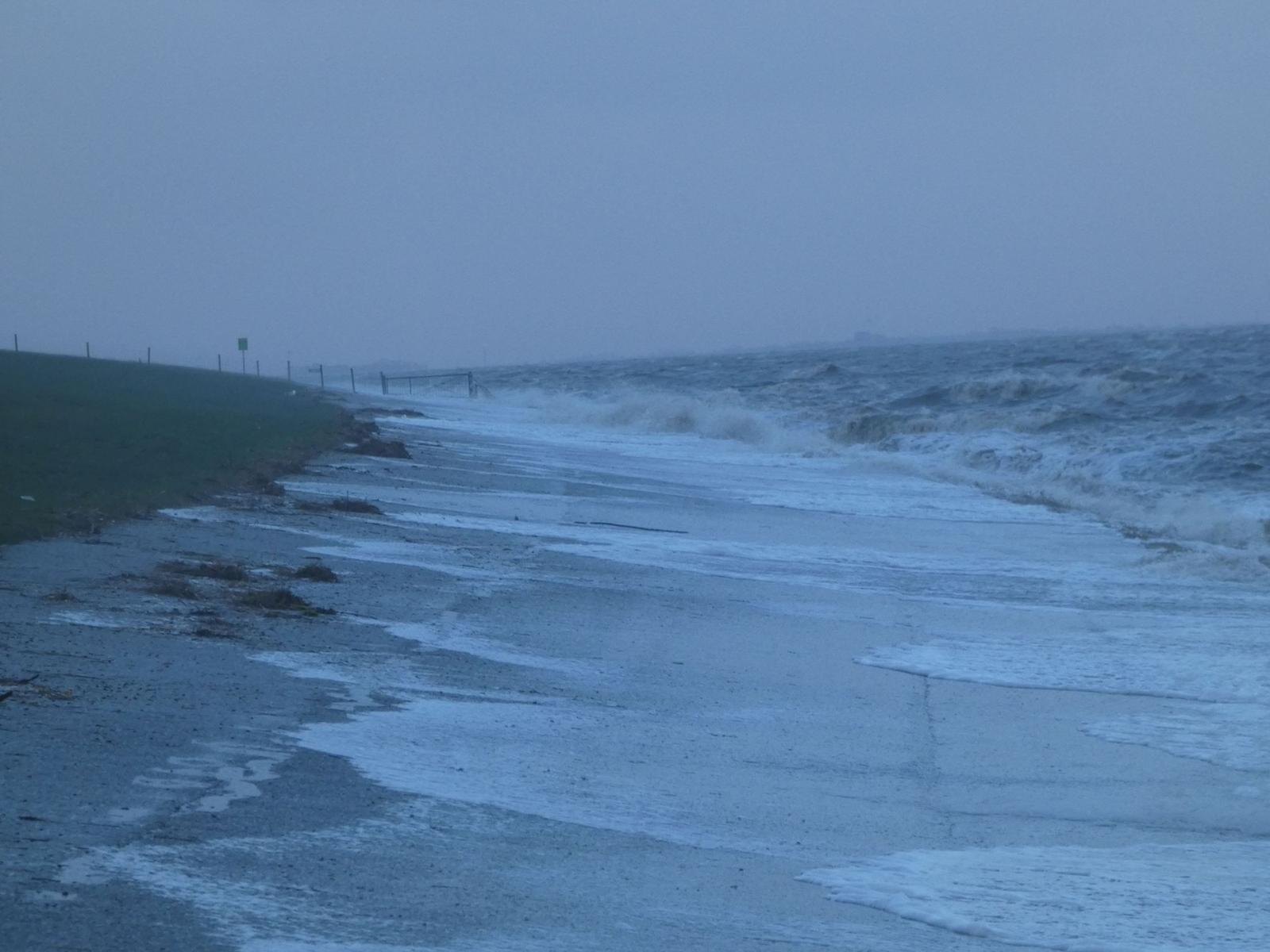 Strand mit Wellen, die an den Uferbereich schlagen, und grasbewachsener Hang im Hintergrund.