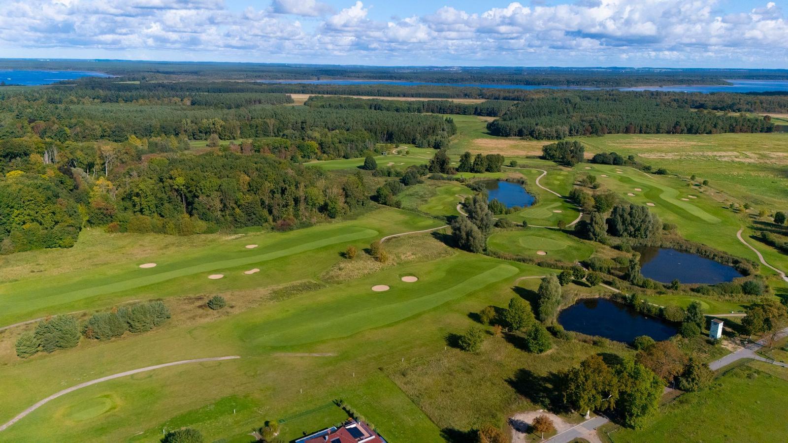 Luftbild eines Golfplatzes mit grünen Fairways, Bäumen und Teichen. Im Hintergrund sind Wälder und ein See sichtbar.