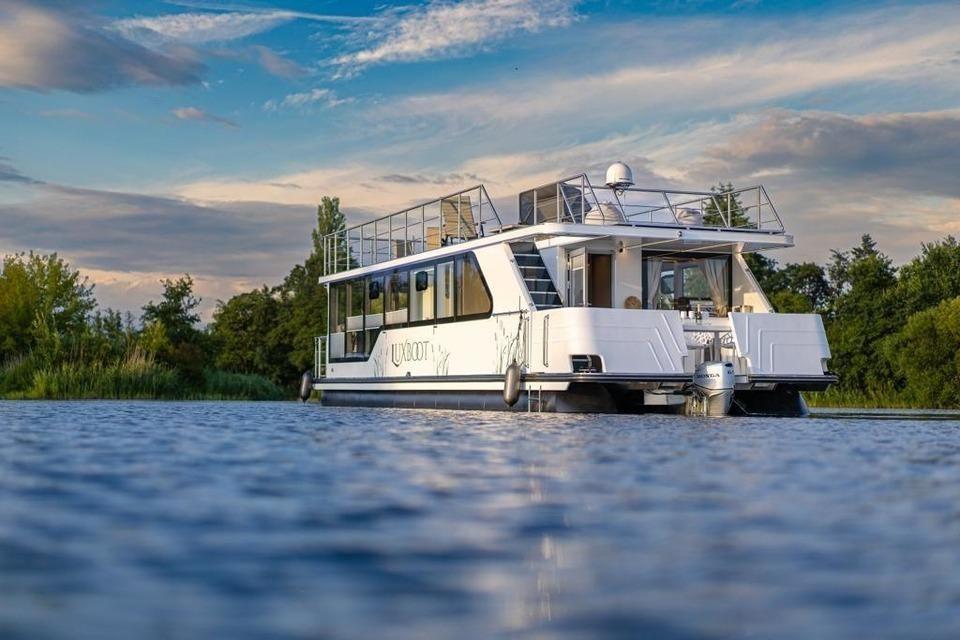 Ein weißes Hausboot mit Balkon liegt auf ruhigem Wasser zwischen Bäumen.