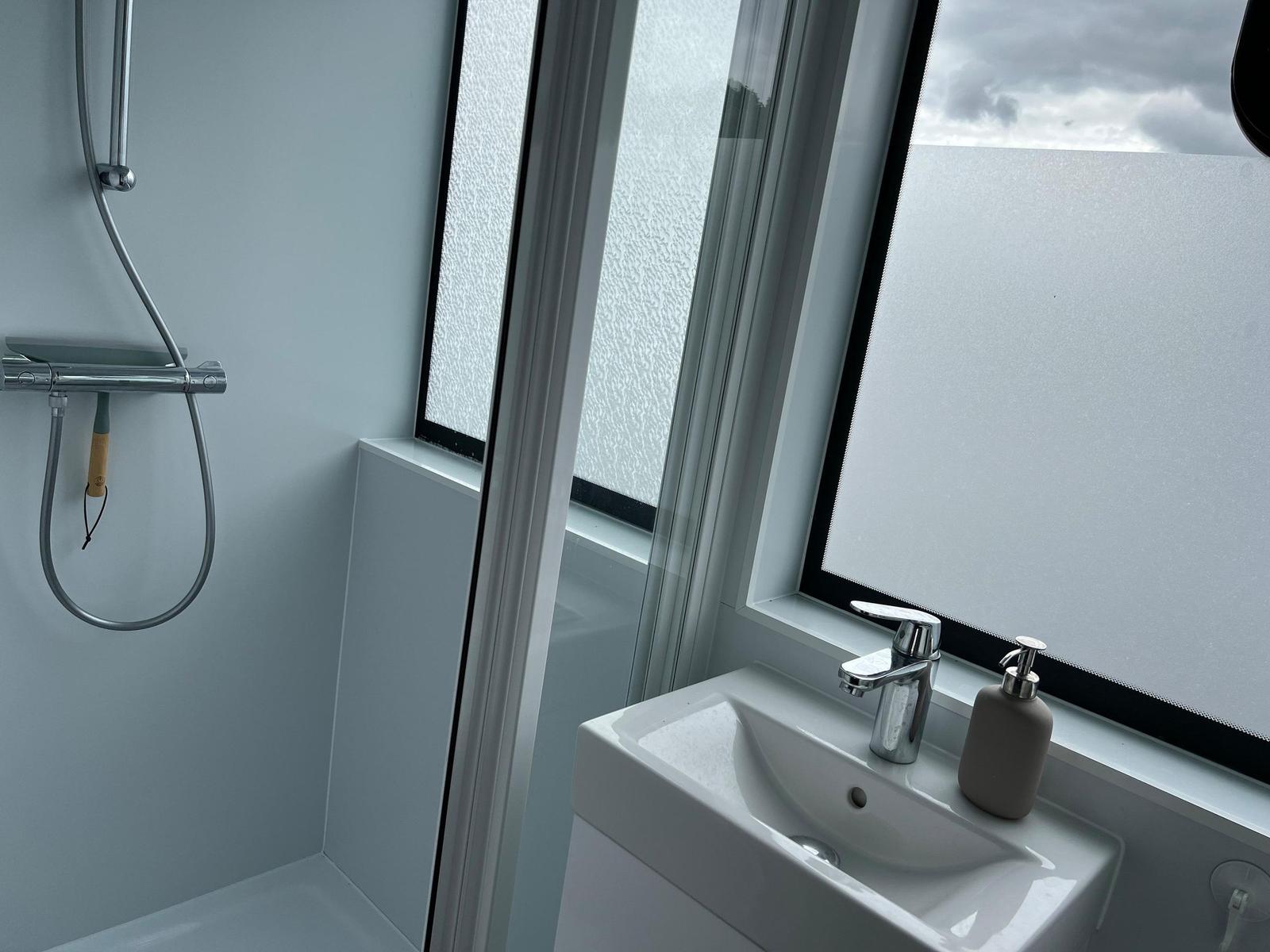 Badezimmer mit Dusche, Waschbecken und Fenster mit getöntem Glas.