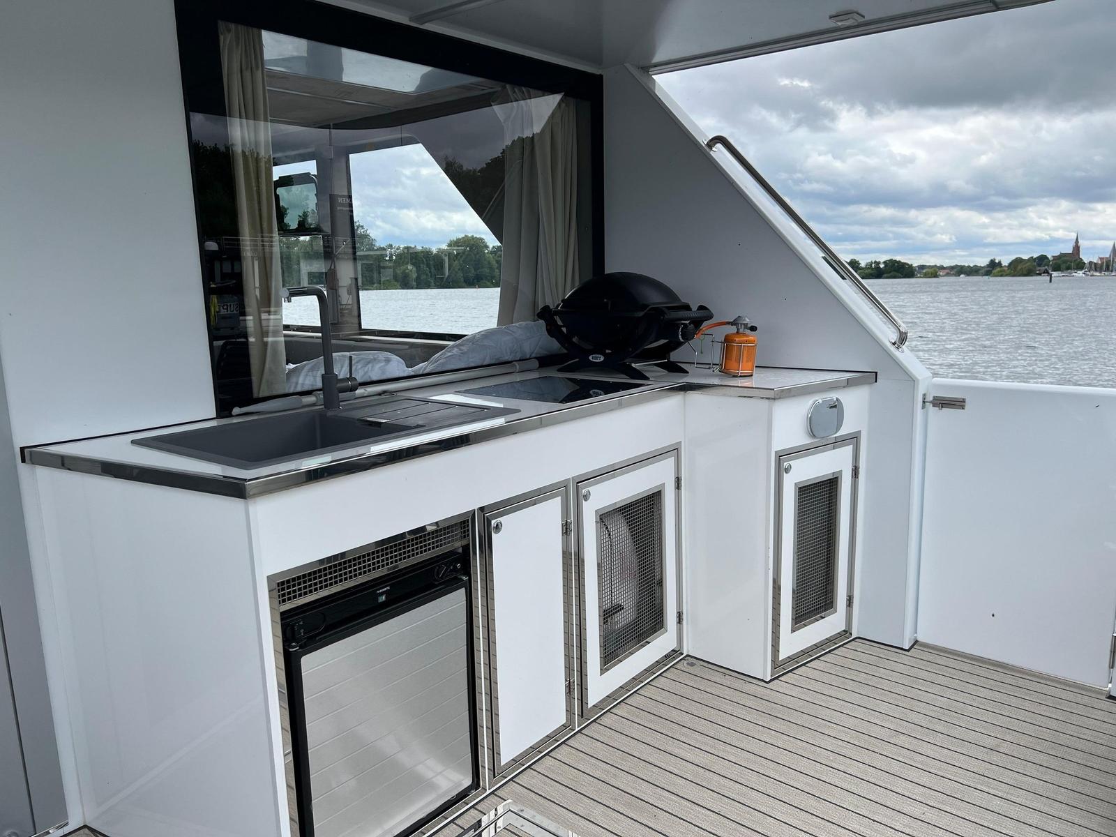 Außenküche mit Spüle, Grill und Kühlschrank an Bord. Blick auf Wasser und Ufer.