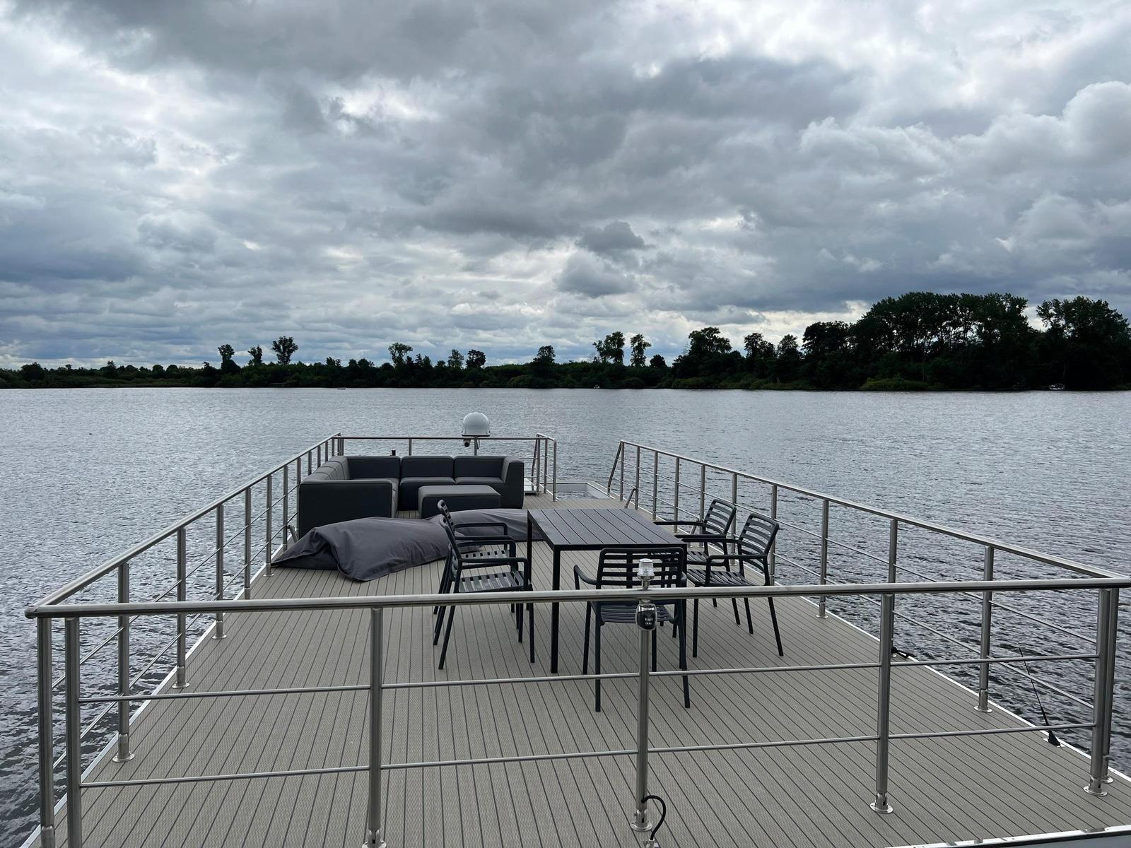 Terrasse mit Sitzgruppe und Tisch über Wasser. Himmel bewölkt, Ufer im Hintergrund.