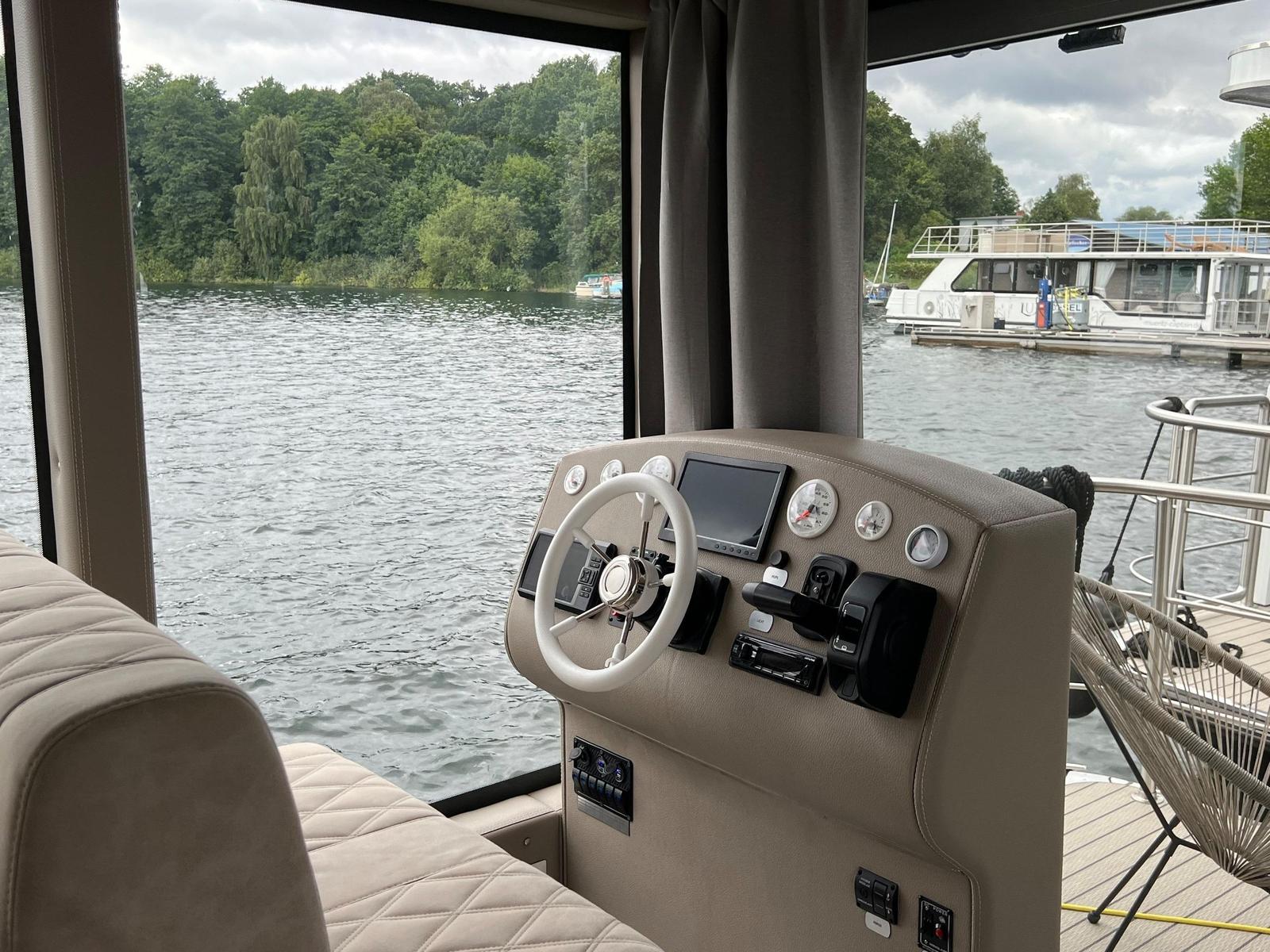 Steuerstand mit Lenkrad und Instrumententafel an einem See mit Blick auf einen Bootssteg.