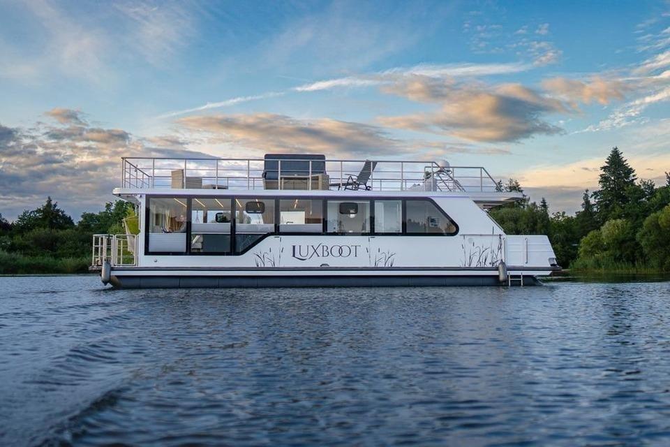 Ein weißes Hausboot mit großem Deck und Fenstern liegt auf ruhigem Wasser.