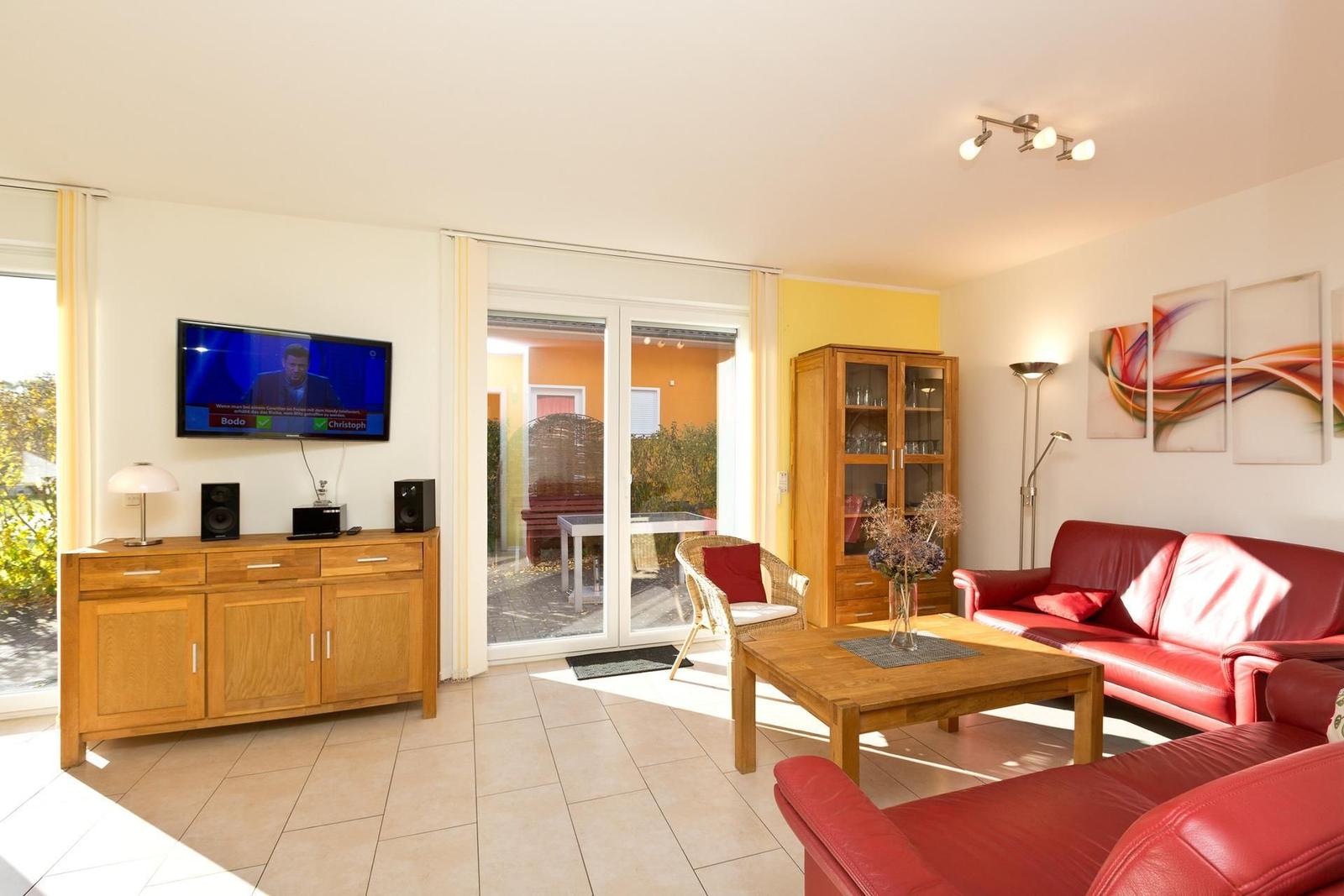 Wohnzimmer mit rotem Sofa, Holzmöbeln und Fernseher. Durchgang zur Terrasse.