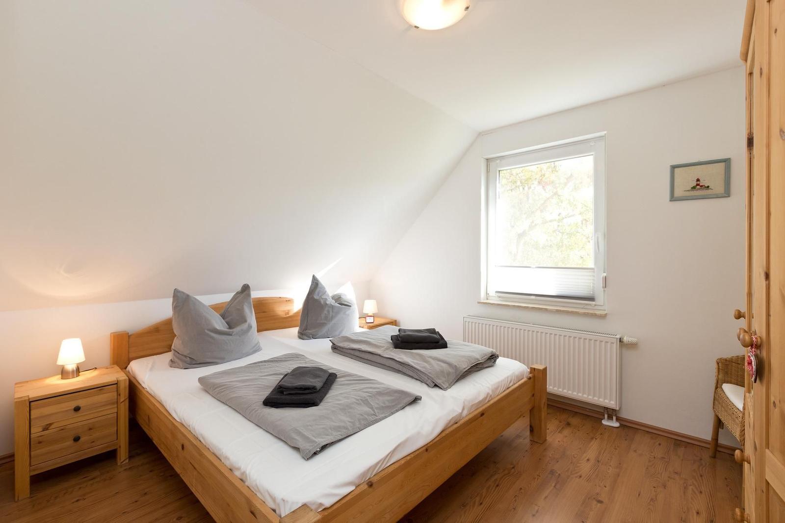 Schlafzimmer mit Holzbett, Nachttisch und Fenster. Holzfußboden und weiße Wände.