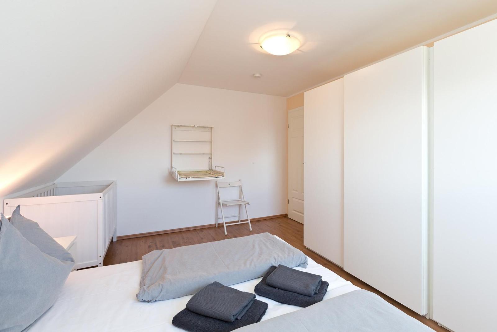 Schlafzimmer mit Babybett, Bett und Kleiderschrank. Wandregal und Stuhl sind vorhanden.