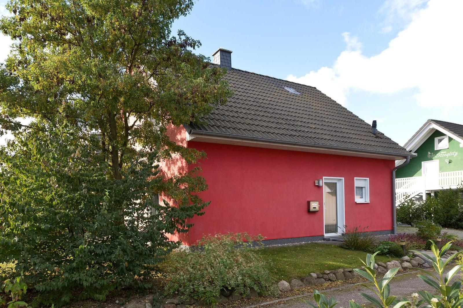 Rotes Haus mit Dach und Eingang. Umgeben von Grünflächen und Bäumen.