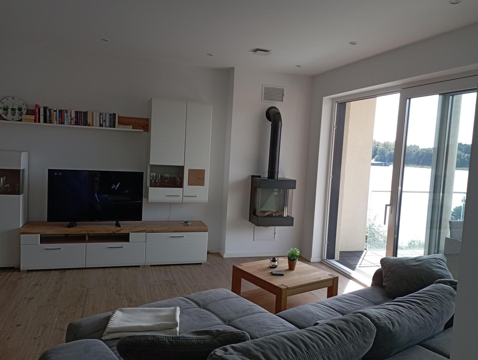 Wohnzimmer mit Sofa, Fernseher und Kaminofen. Durchgang zur Terrasse mit Blick auf Wasser.