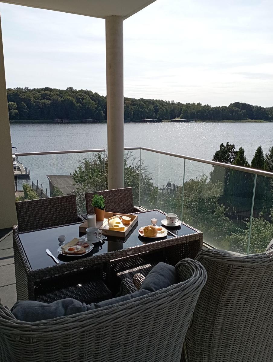 Terrasse mit Tisch und Stühlen, Blick auf einen See mit Wald im Hintergrund.