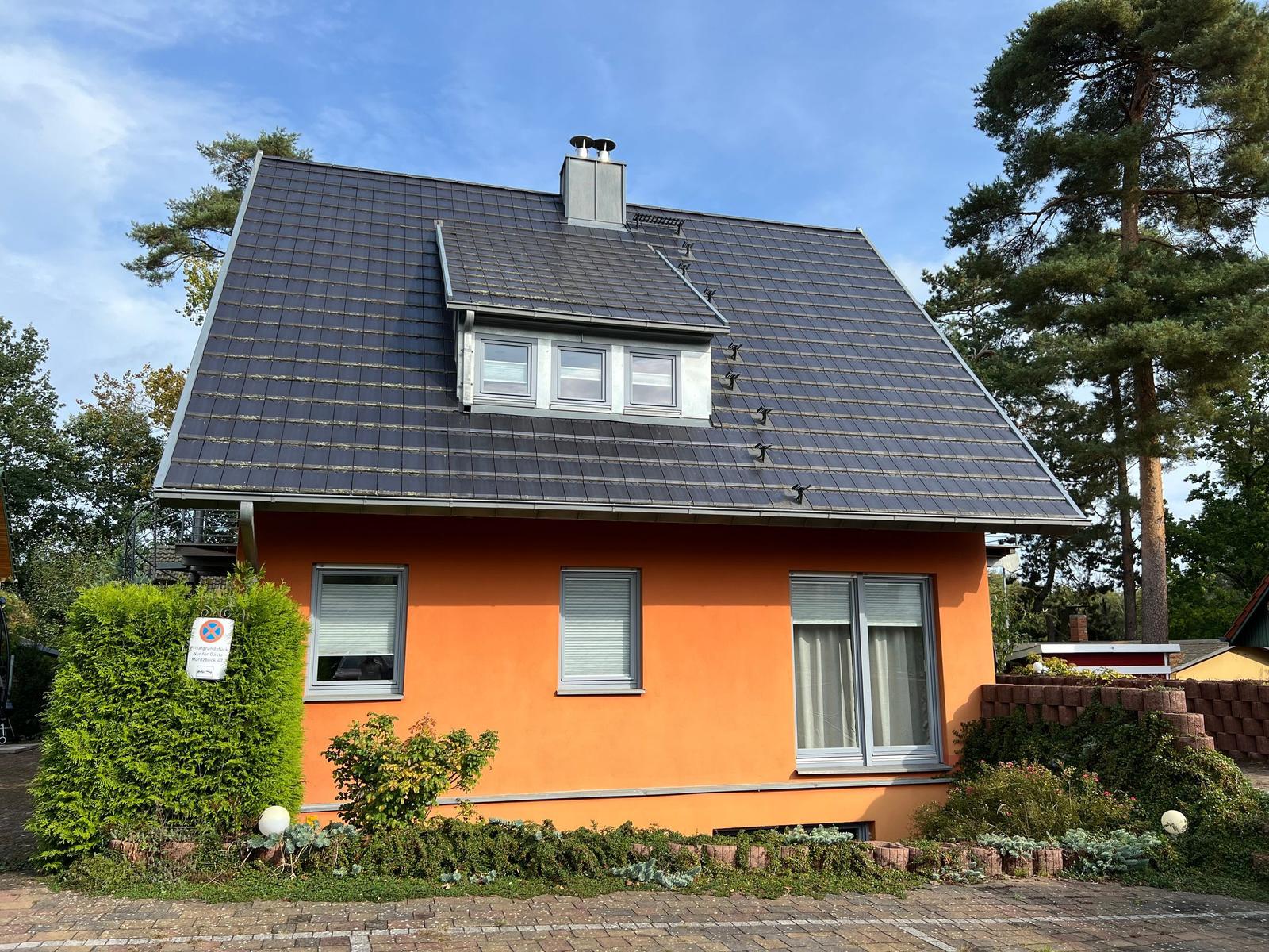 Ein orange gestrichenes Haus mit dunklem Dach und Fenstern. Umgeben von Bäumen und einem gepflasterten Weg.