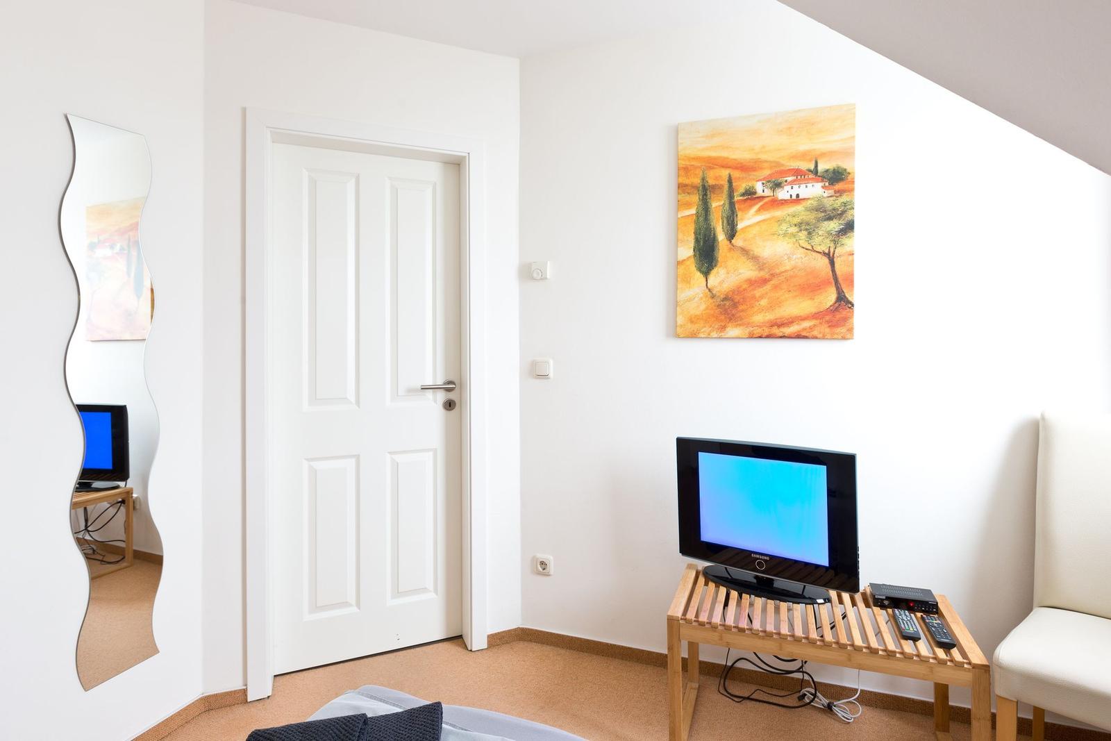 Ein Raum mit Fernseher, Spiegel und Wandbild. Tür und Stuhl sind sichtbar.