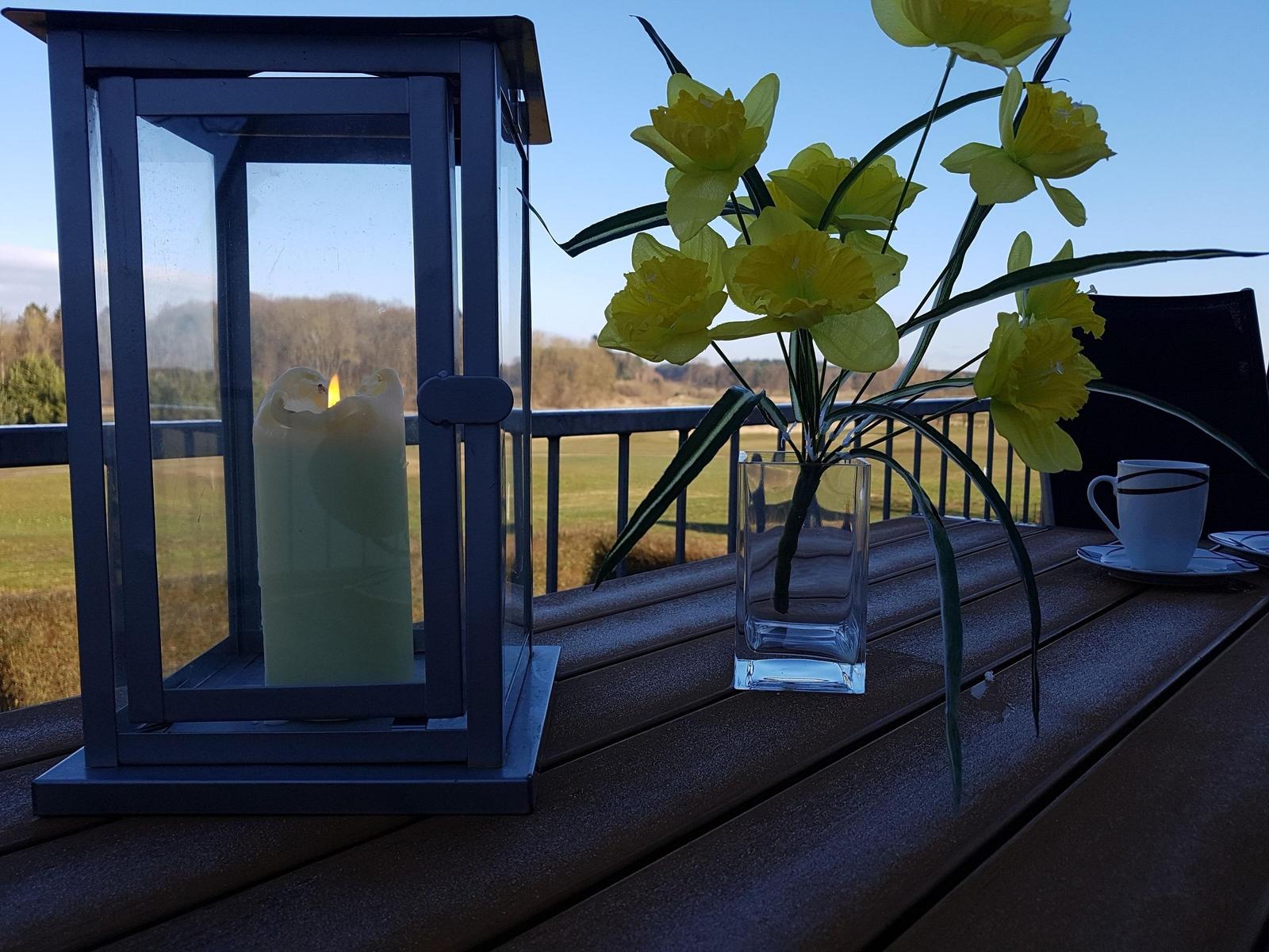 Auf der Terrasse steht eine Laterne mit Kerze, eine Vase mit Narzissen und eine Tasse.