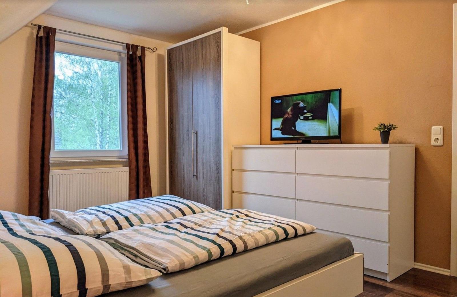 Schlafzimmer mit Bett, Kleiderschrank und Fernseher auf Schrank. Fenster mit Blick auf Bäume.
