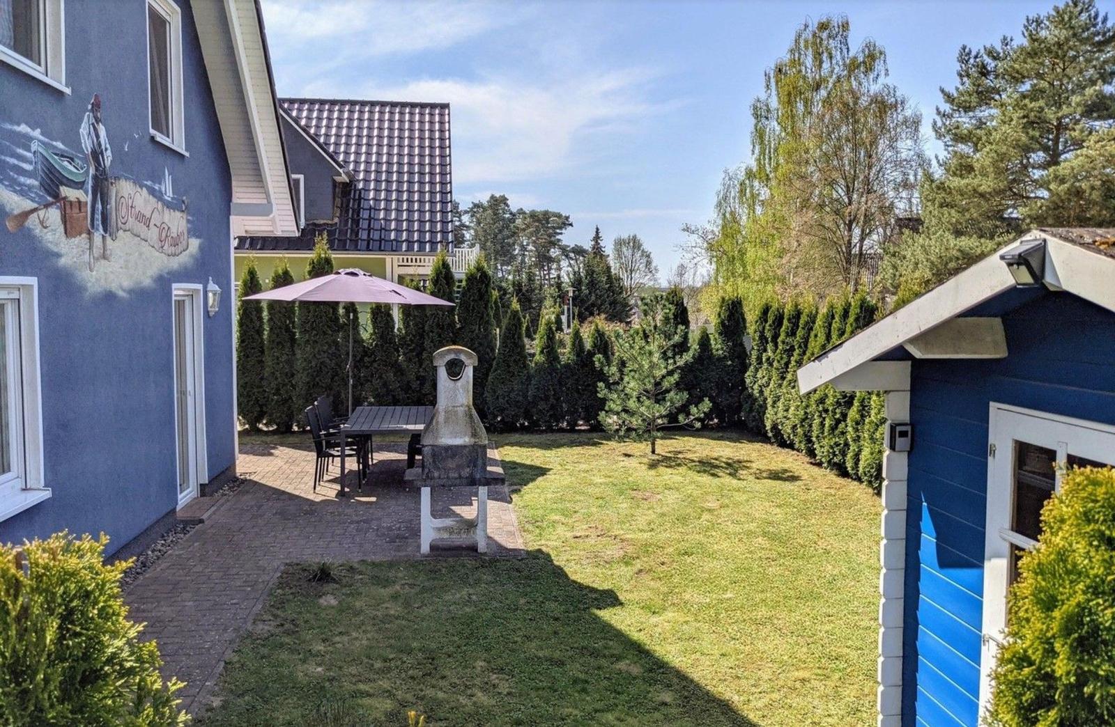 Hinterhof mit Grill, Tisch, Stühlen und Sonnenschirm. Grünfläche und Hecke umgeben den Raum.
