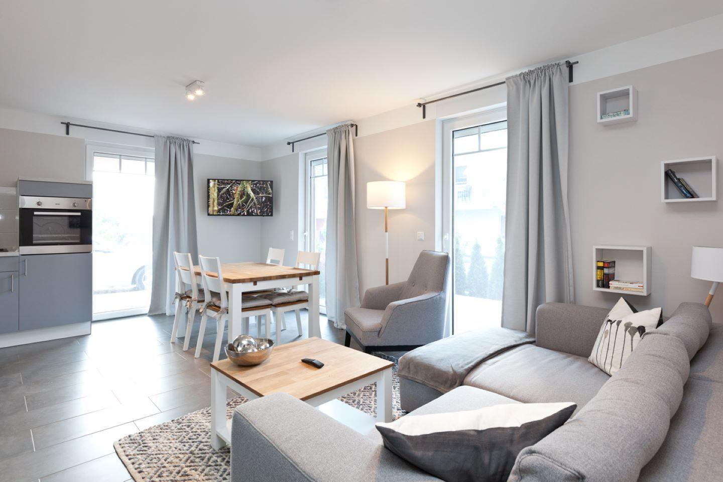 Wohn- und Essbereich mit Sofa, Esstisch und Küchenzeile. Große Fenster und Türen.