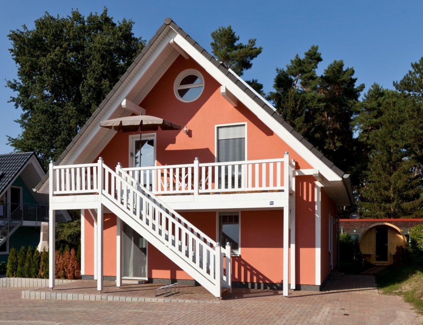 Ein zweistöckiges Haus mit Balkon und Außentreppe. Umgeben von Bäumen und einem gepflasterten Hof.