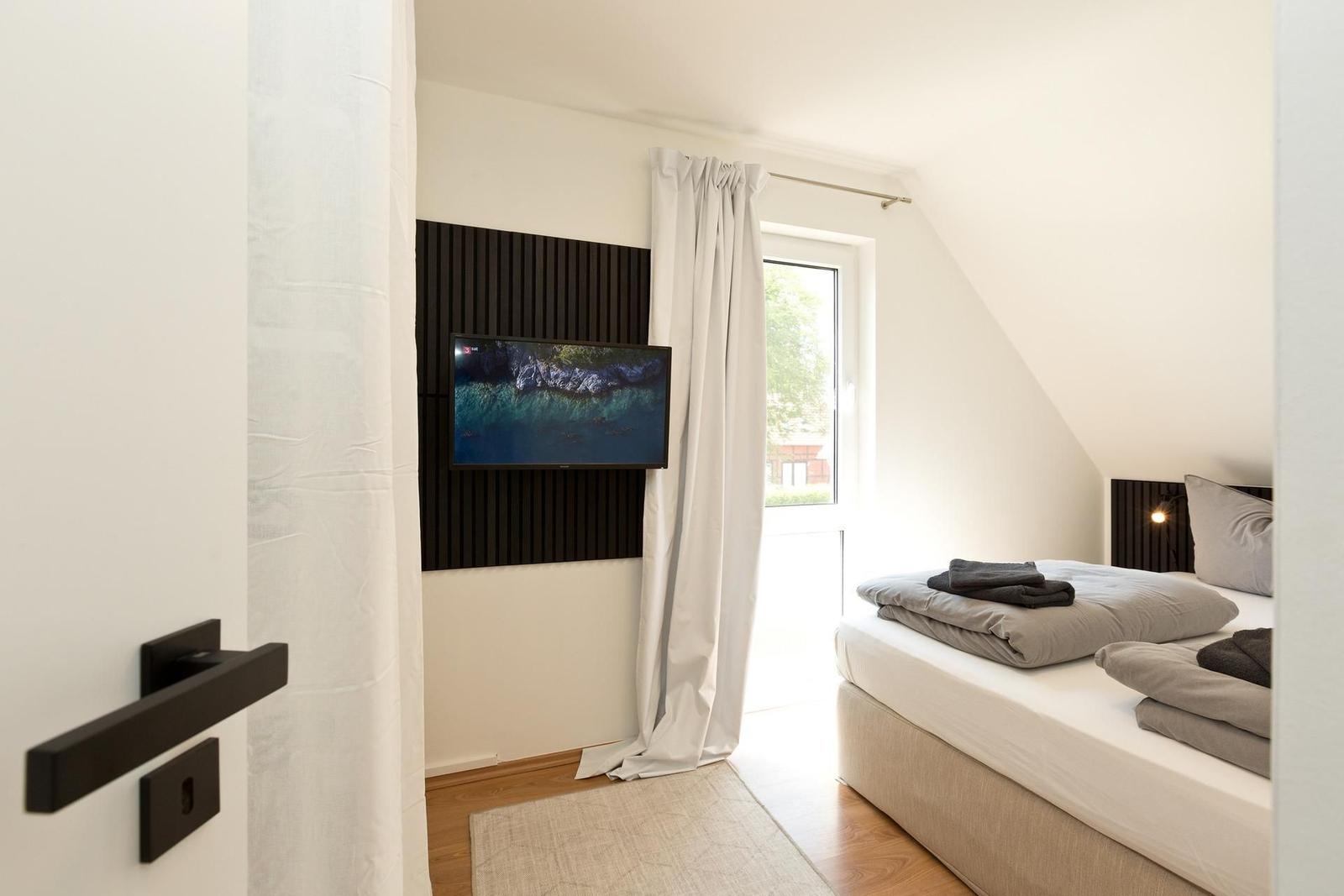 Schlafzimmer mit Bett, TV und Fenster. Holzboden und weiße Wände.