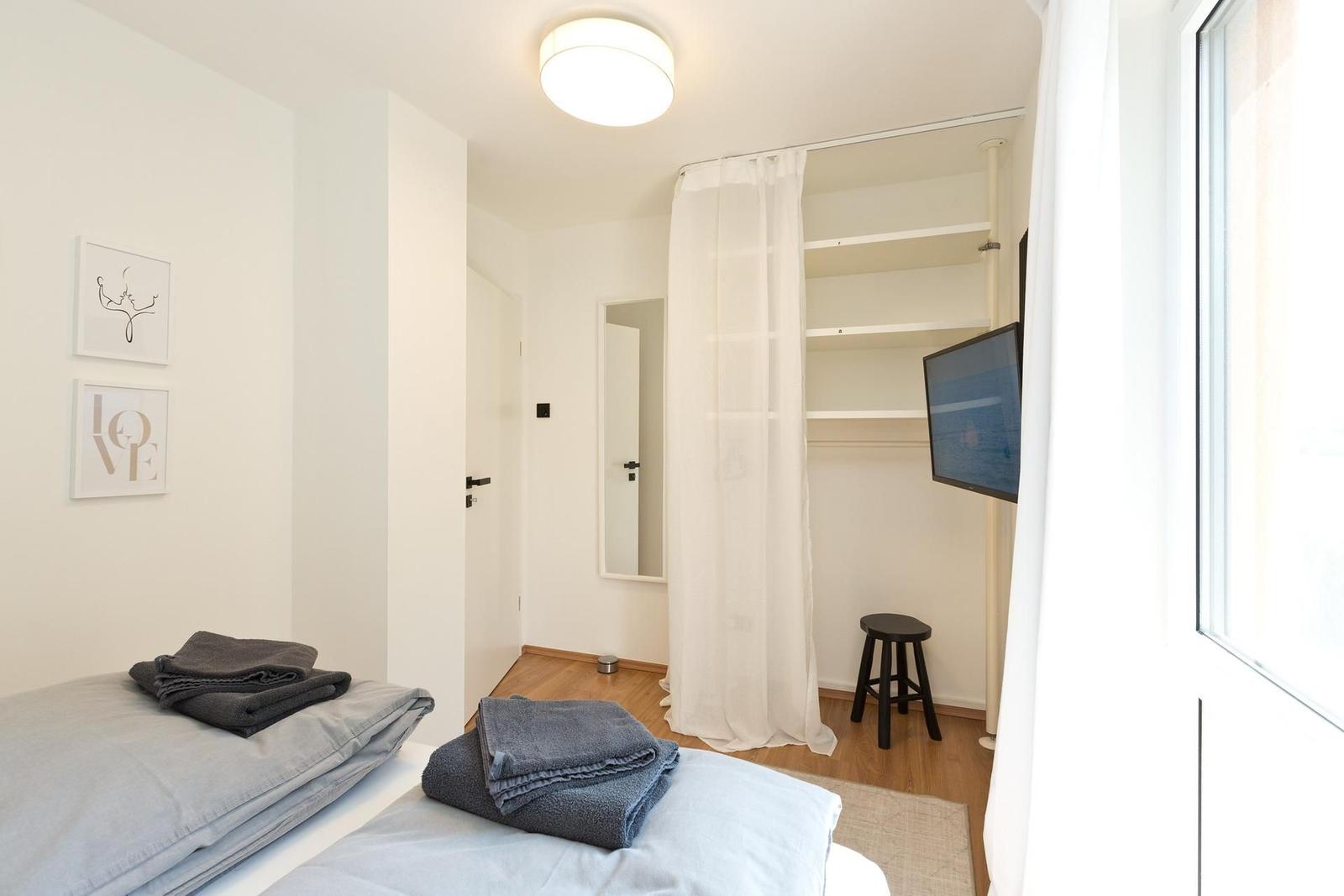 Schlafzimmer mit Bett, Kleiderschrank und Fernseher. Fenster mit weißen Vorhängen.