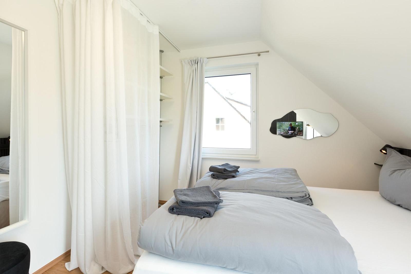 Schlafzimmer mit Dachschrägen, Fenster, Spiegel und Bett mit grauer Bettwäsche.