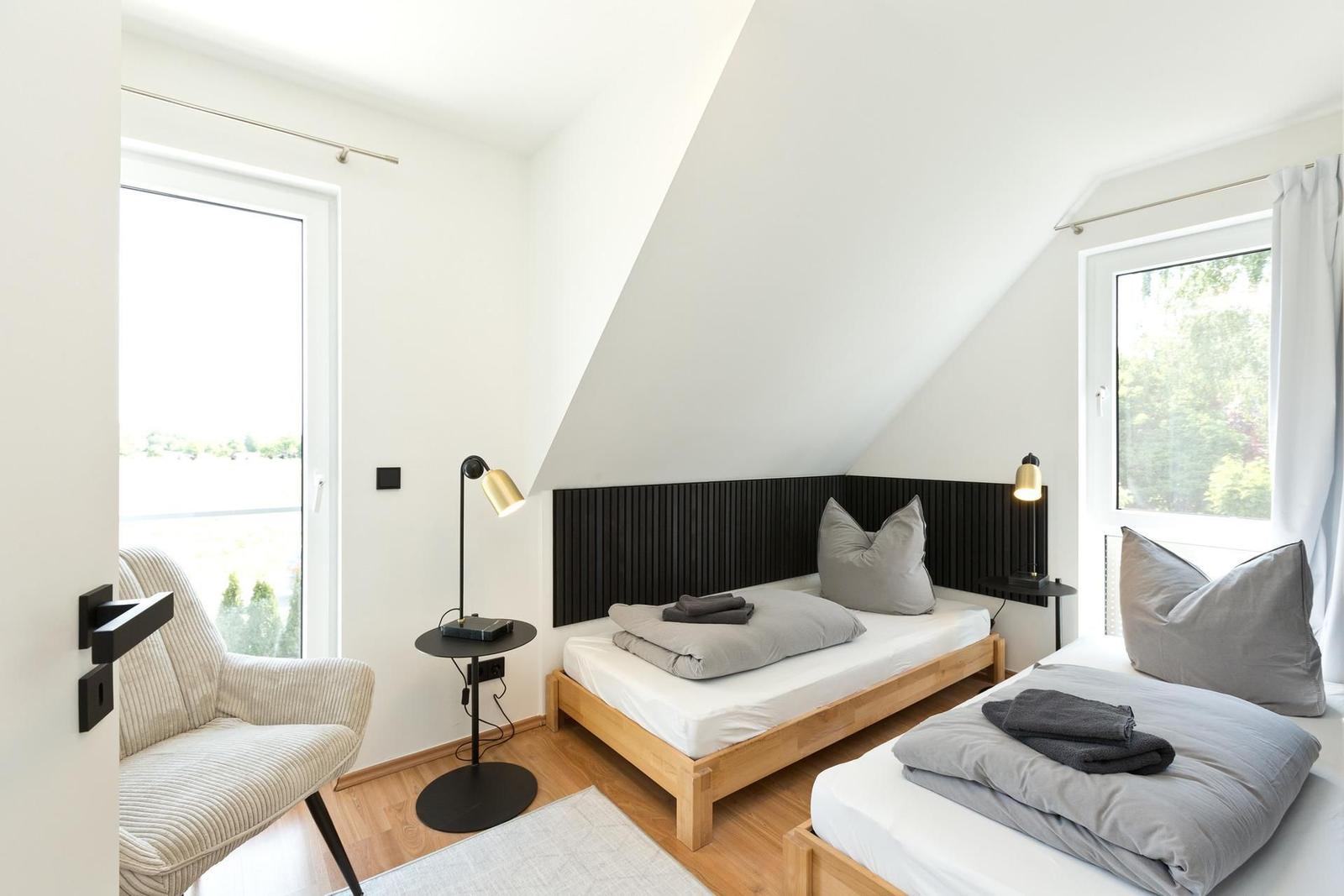 Zwei Betten mit Holzrahmen, schwarze Kopfteilwand, zwei Fenster und ein Sessel.