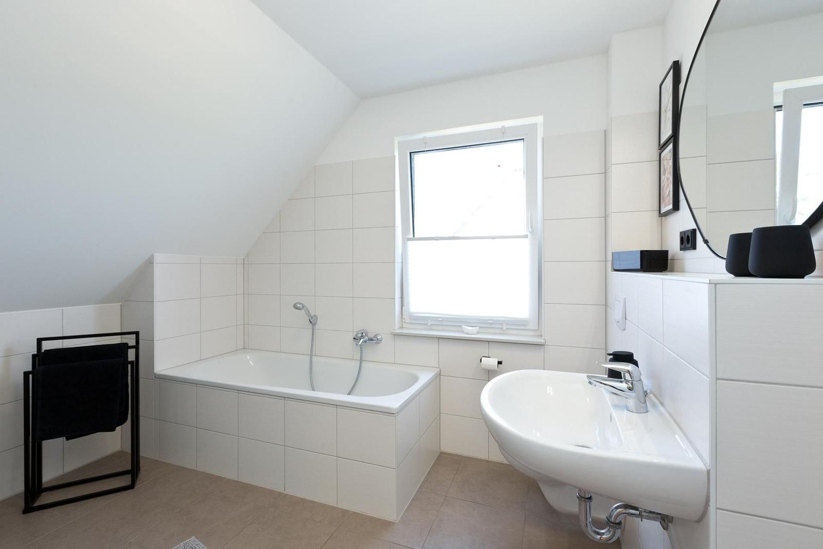Badezimmer mit Badewanne, Waschbecken und Fenster. Weiße Fliesen und schwarze Handtuchhalter.
