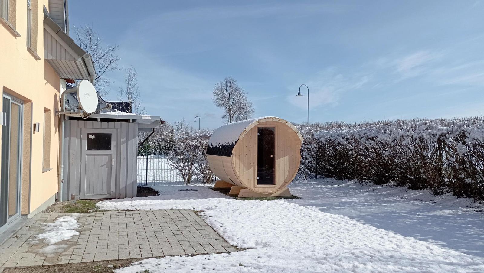 Holzsauna im Schnee vor einem Haus mit Satellitenschüssel und Heizung.
