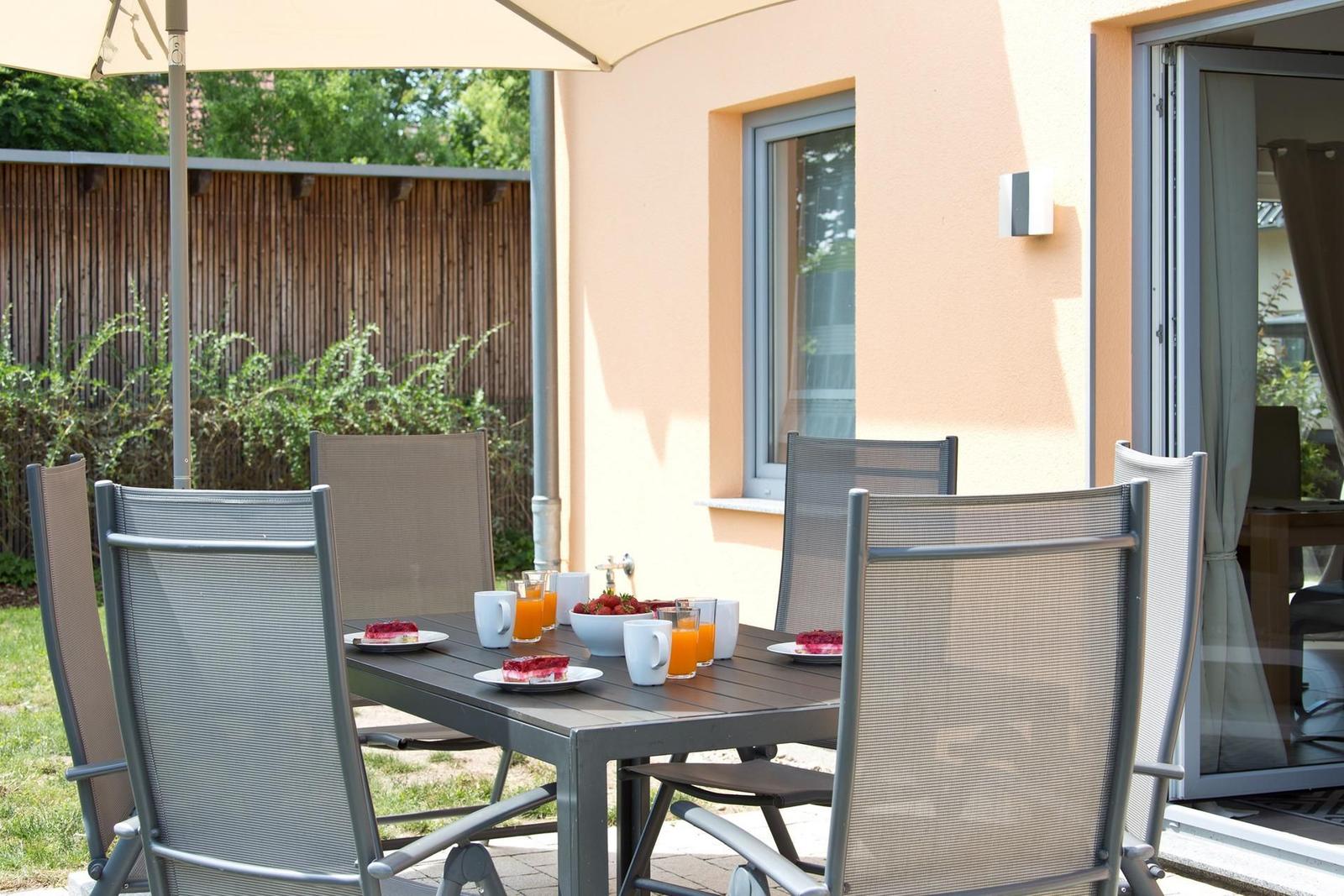 Terrasse mit Esstisch, Stühlen und Sonnenschutz. Auf dem Tisch stehen Tassen und Obst.